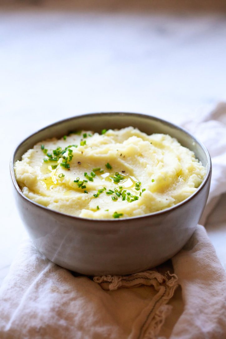 A bowl of mashed cauliflower garnished with chives on a marble countertop. Cauliflower "mashed potatoes" are a creamy and cozy healthier side dish perfect for the holidays or any day. This garlic cauliflower mash recipe is keto and vegan friendly. 