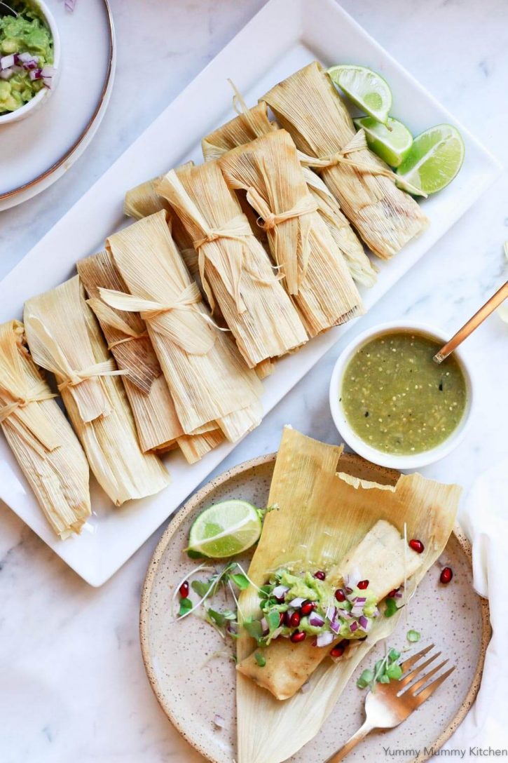 Easy homemade vegetarian or vegan tamales made with Mexican masa, black beans, chilies, and cheese. Beautiful homemade tamales on a platter next to an open tamale on a plate served with salsa verde and guacamole. 