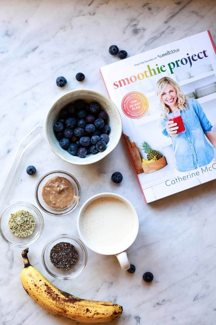 Ingredients for a blueberry banana chia smoothie alongside the smoothie project cookbook on a marble countertop. 