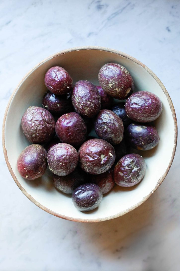 A large bowl filled with wrinkled ripe purple passion fruit sits on a marble countertop. 
