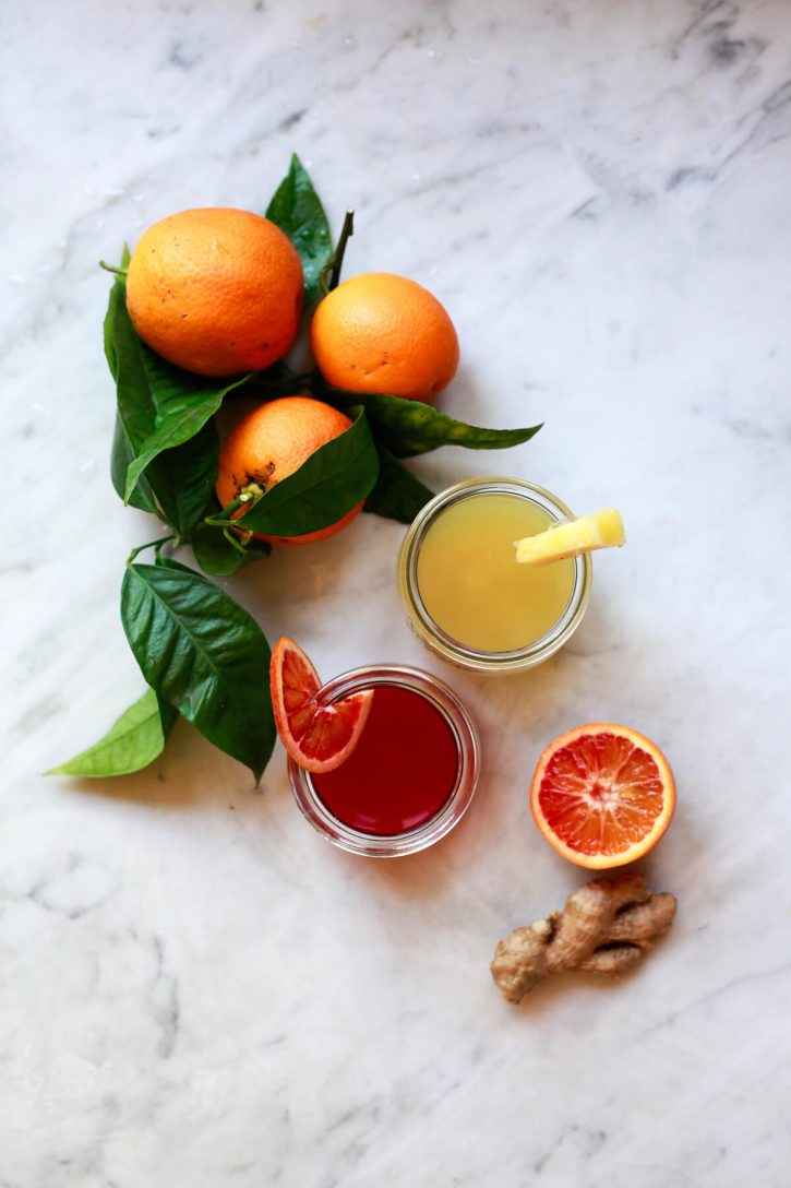 Two wellness shots sit on a marble countertop next to ginger root and oranges. 