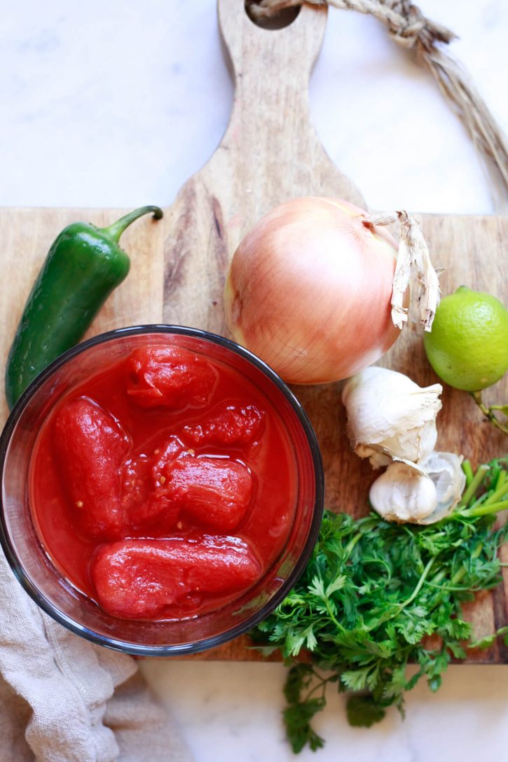 The ingredients for an easy homemade cooked salsa sit on a cutting board: canned whole tomatoes, onion, jalapeno, garlic, cilantro, and lime. 