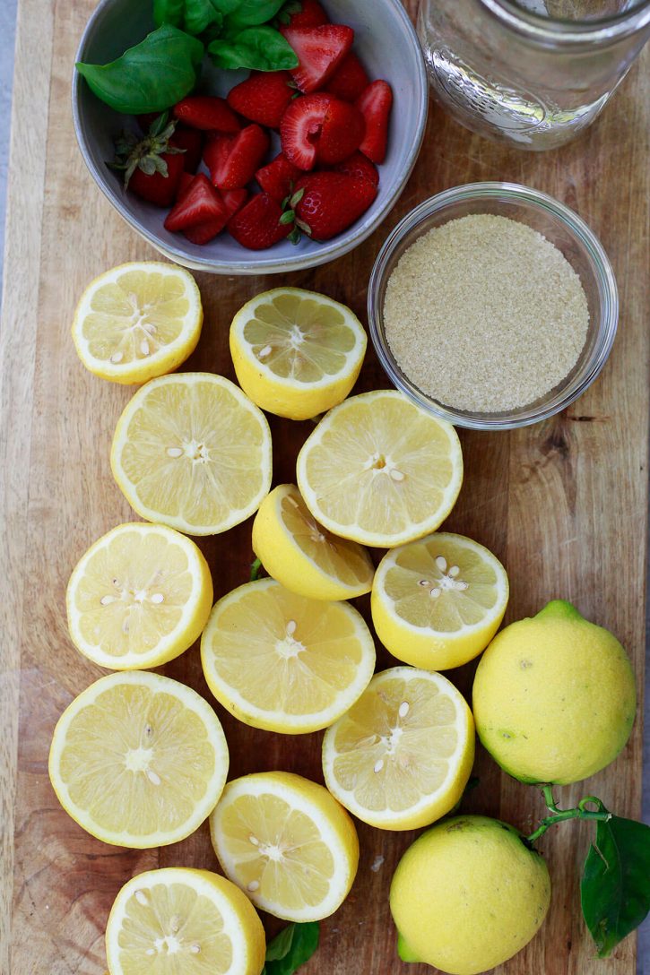 strawberry lemonade ingredients Halved lemons, strawberries, sugar, and water on a cutting board to make homemade strawberry pink lemonade.