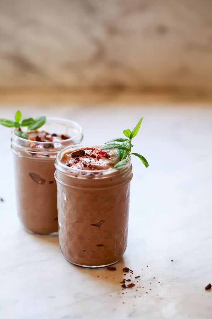Two chocolate avocado smoothies garnished with mint and dark chocolate sit on a marble kitchen countertop. 