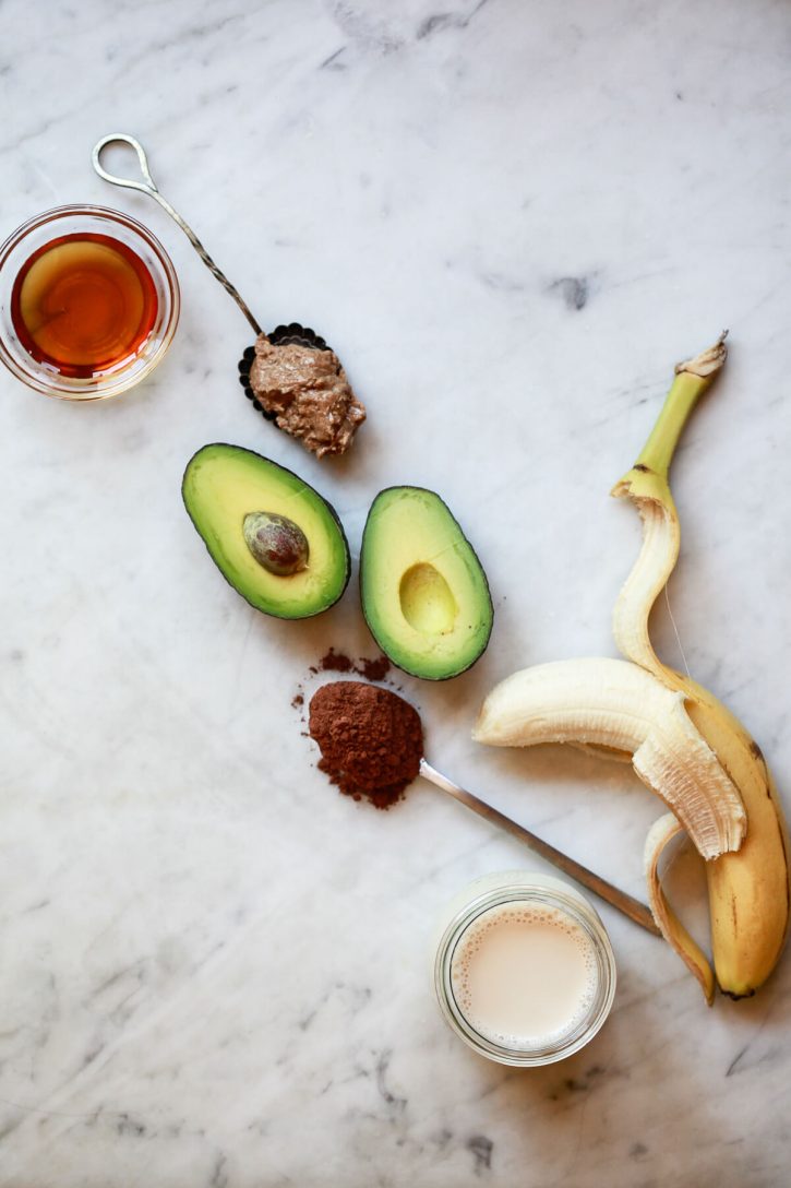 The ingredients for a chocolate banana avocado smoothie on a marble kitchen counter: banana, avocado, cacao powder, almond milk, almond butter, and maple syrup. 