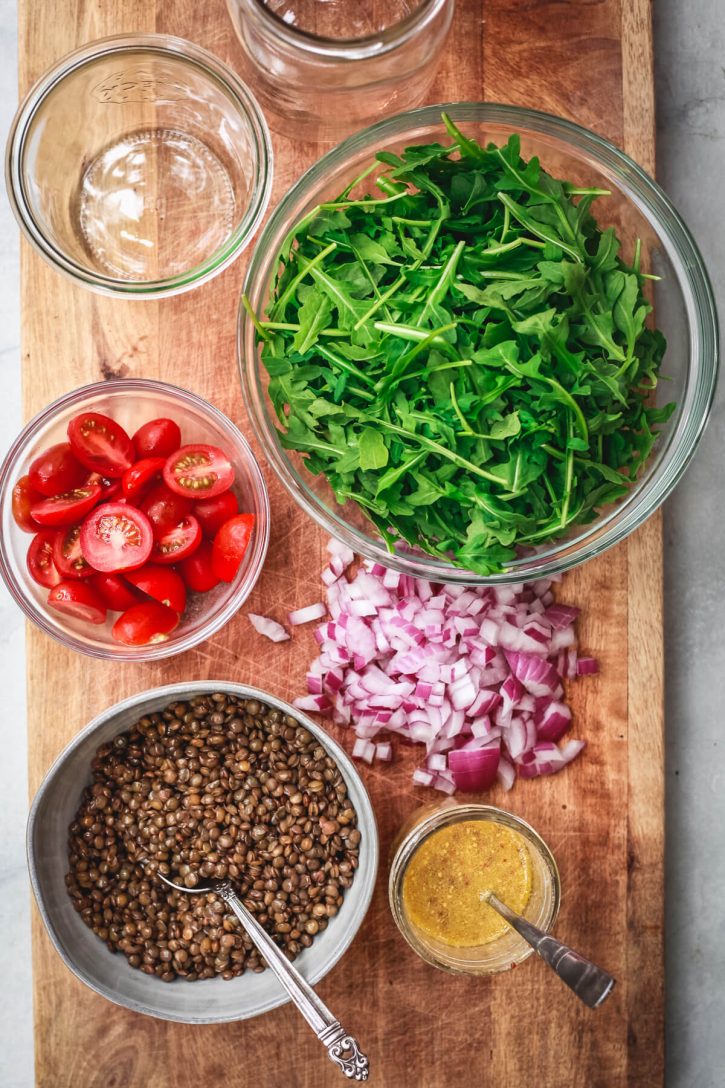 Cold French lentil salad ingredients sit on a cutting board to assemble into mason jars. Ingredients shown include cooked French lentils, halved cherry tomatoes, arugula, diced red onion, and a prepared homemade mustard vinaigrette. 