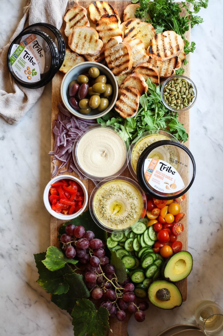 A beautiful charcuterie board topped with vegan ingredients like grilled crostini bread, hummus, peppers, cucumber, tomatoes, avocado, and arugula. 