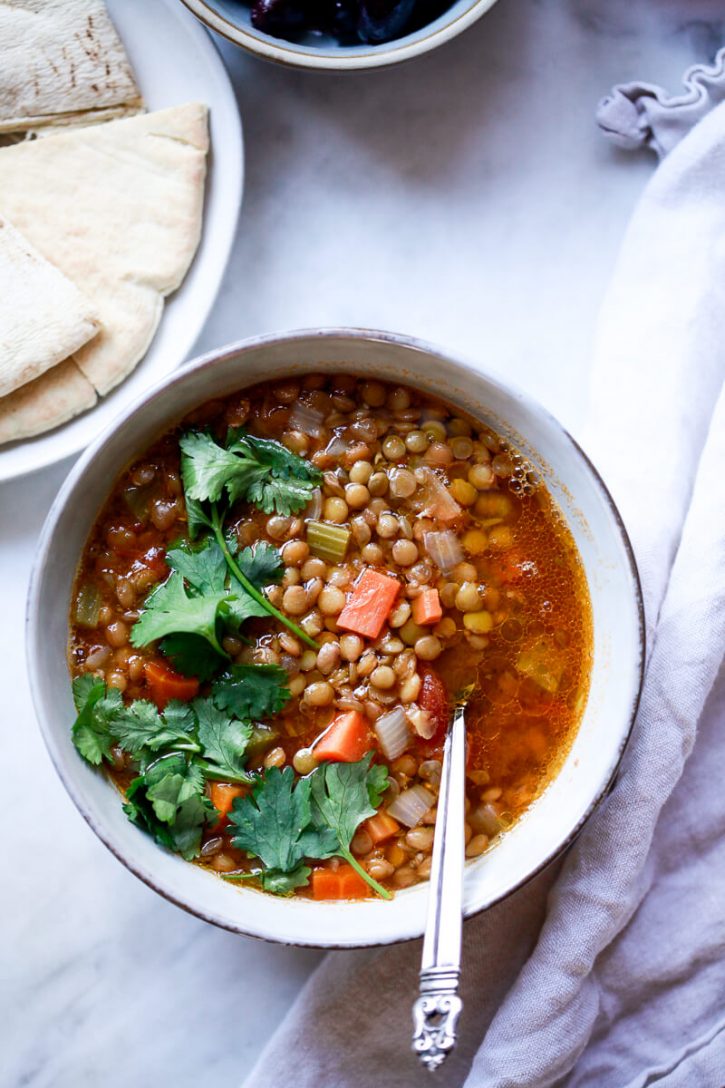 A bowl of vegan traditional Greek lentil soup garnished with cilantro sits on a marble countertop next to pita bread and olives. 