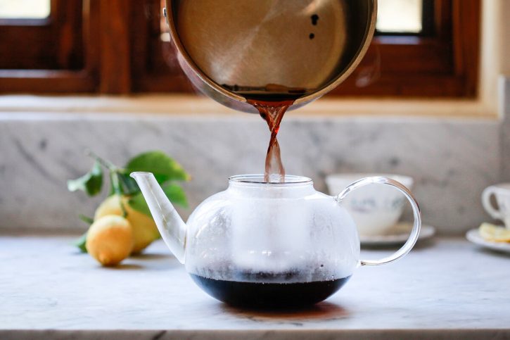 A saucepan of homemade elderberry tea is poured through a strainer in a teapot. 