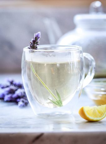 A cup of lavender tea in a glass cup garnished with a sprig of fresh lavender.