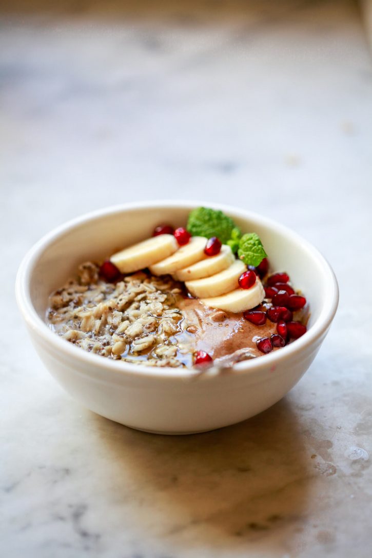 Photograph of a white bowl filled with chia seed protein oatmeal topped with sliced banana, pomegranate seeds, and almond butter. 