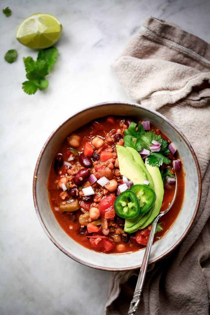Overhead photo of the best vegan chili ever topped with avocado, jalapeno, and cilantro in a ceramic bowl. This high protein vegan chili has 3 types of beans visible (chickpeas, kidney beans, and black beans). 