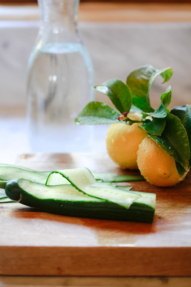 A jug of water sits behind an English cucumber that is getting peeled into ribbons on a cutting board with lemons to the side. 