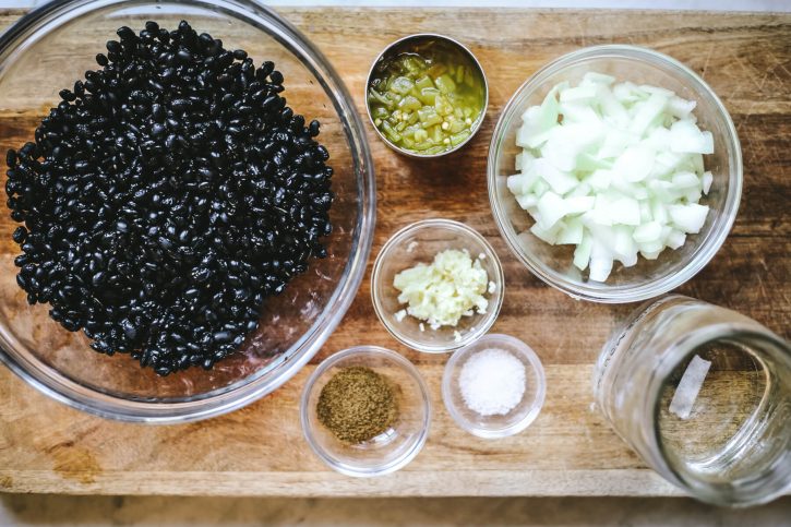 An overhead photograph of dry balck beans, cumin, garlic, salt, onion, chilies, and water. These are the ingredients for instant pot black beans. 