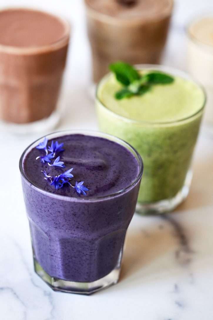 5 beautiful and delicious protein shakes on a kitchen counter with a purple shake topped with edible flowers in the foreground. 