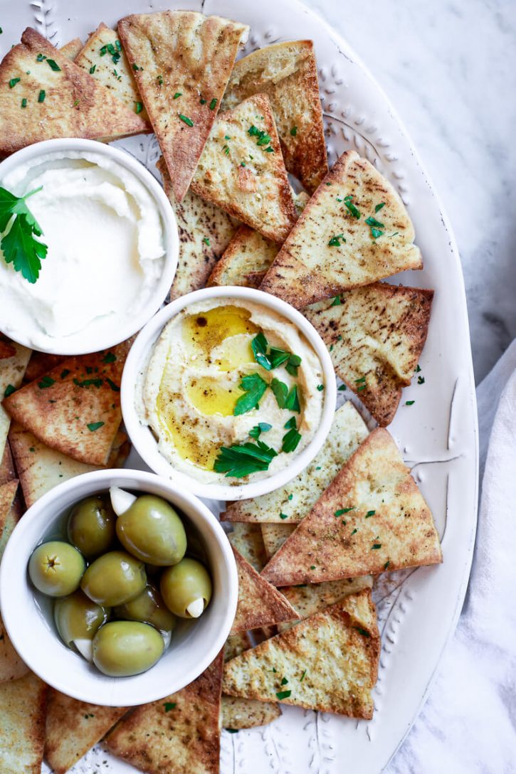 A white platter filled with homemade baked or air fried pita chips, hummus, labneh, and olives. An easy and healthy pita chip recipe. 