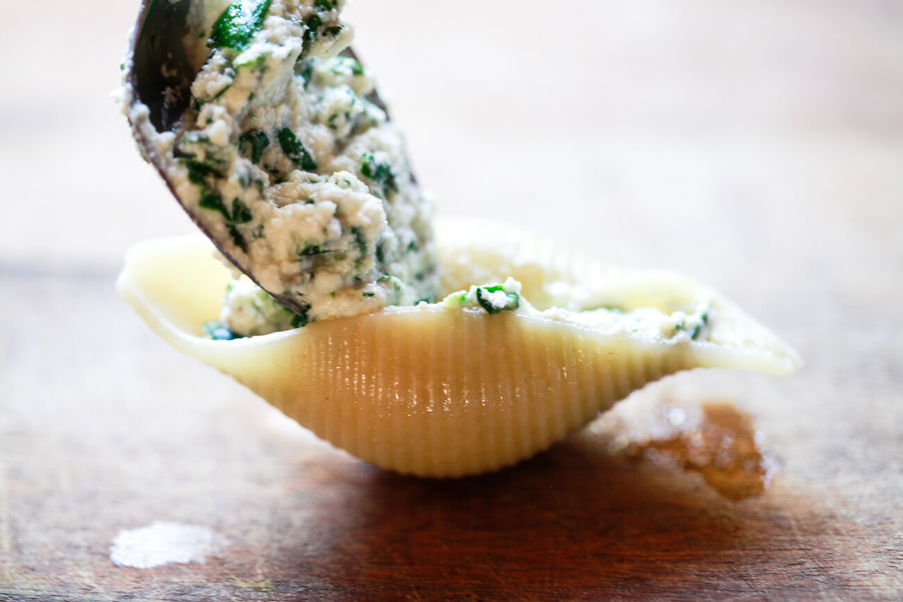 A close-up photo of a spoon filling a cooked Conchiglioni pasta shell with spinach ricotta filling. 