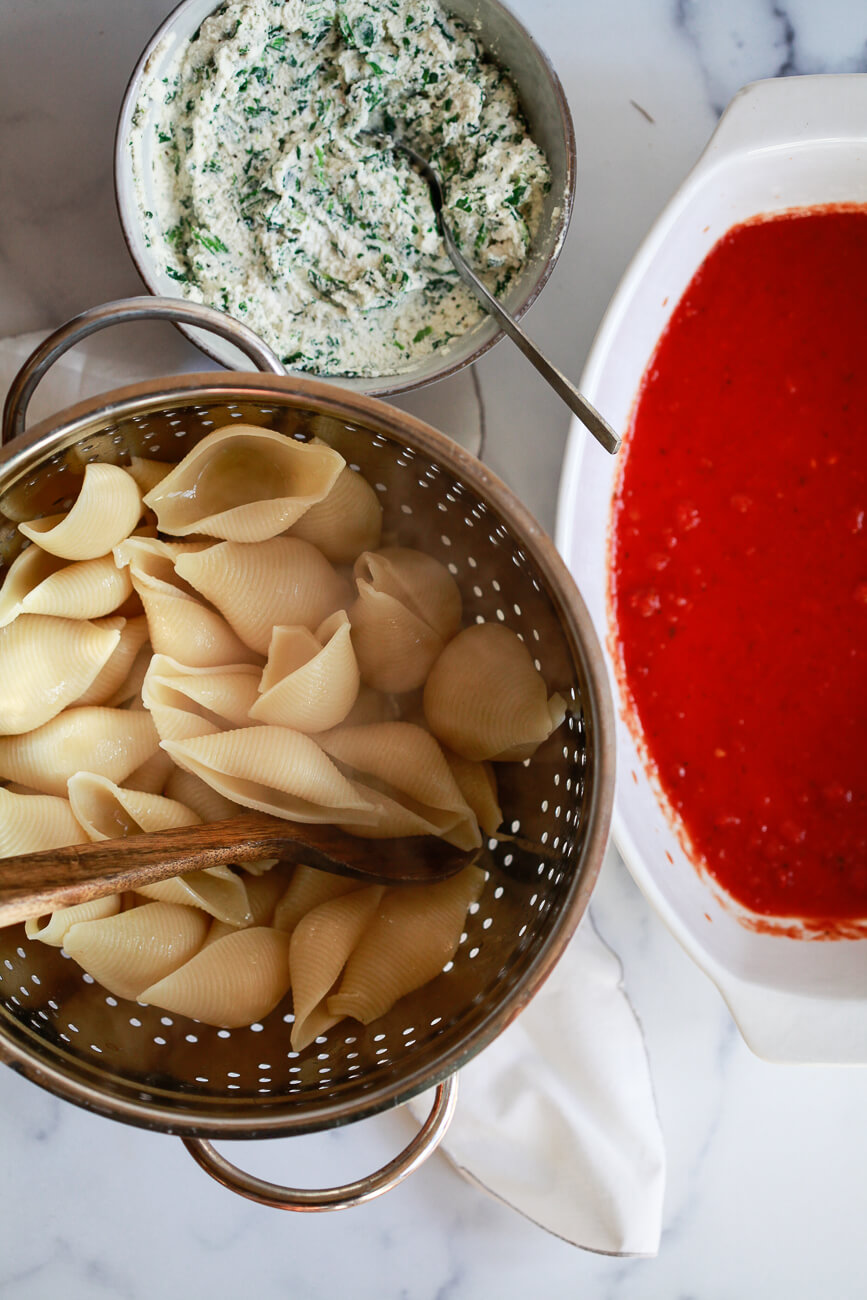 An overhead photo of cooked shell pasta in a colander, a bowl of ricotta filling, and a casserole dish filled with marinara sauce. 