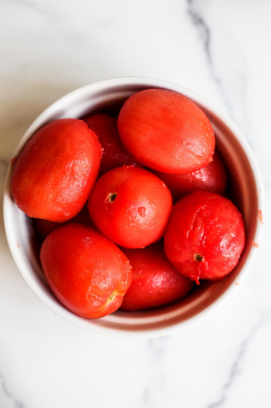 An overhead photo of a bowl of blanched peeled tomatoes on a marble countertop. 