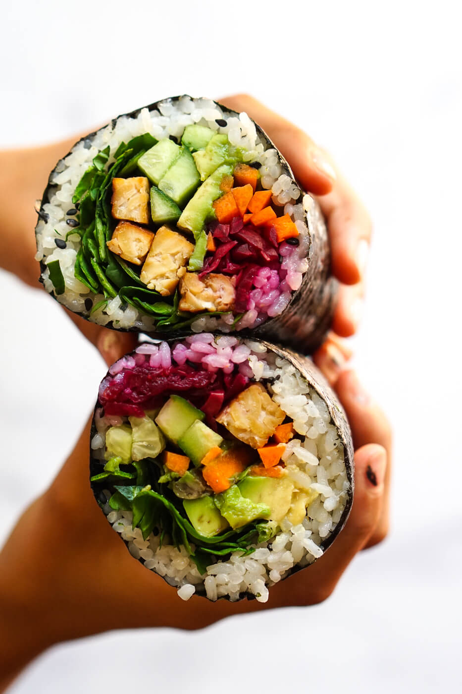 Photograph of hands holding two halves of a sushi burrito filled with vegetables and tempeh. 