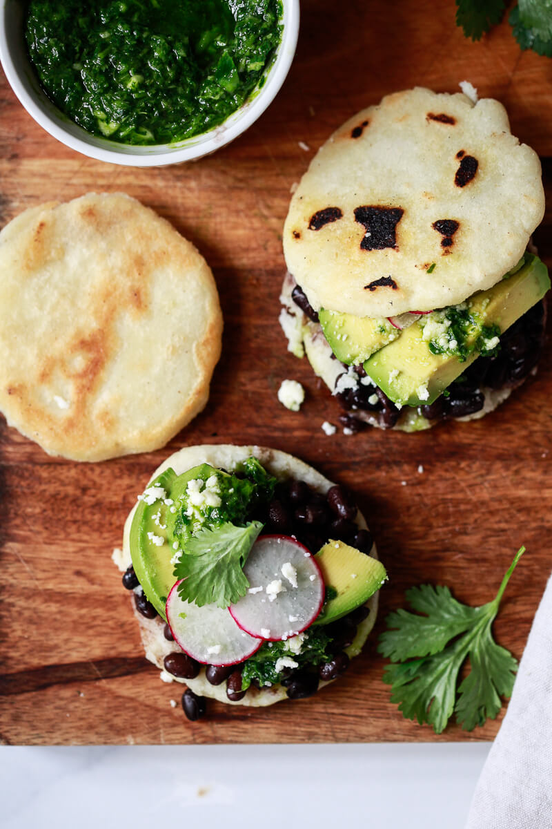Homemade Arepas Venezolanas topped with black beans and avocado on a cutting board with a bowl of chimichurri sauce on the side. 