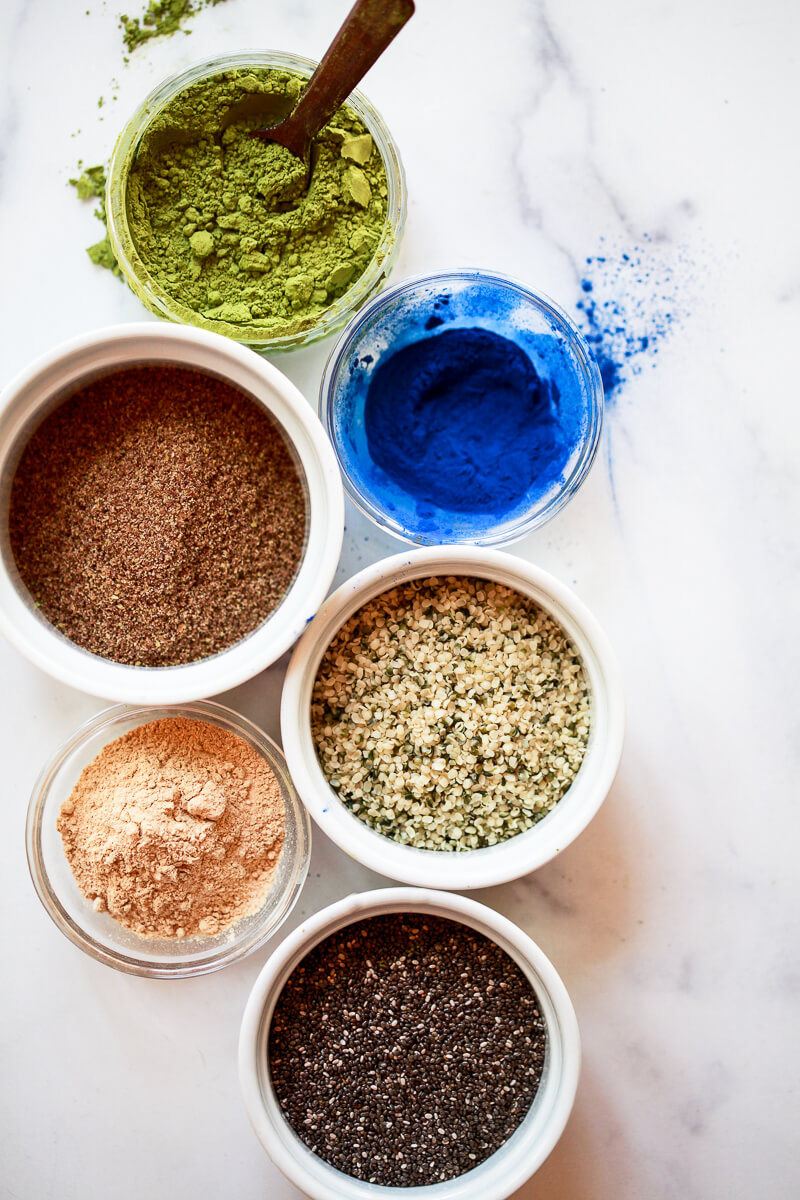 An overhead photo of superfood smoothie supplements. These include matcha powder, blue spirulina, flax, hemp, maca, and chia seeds. 