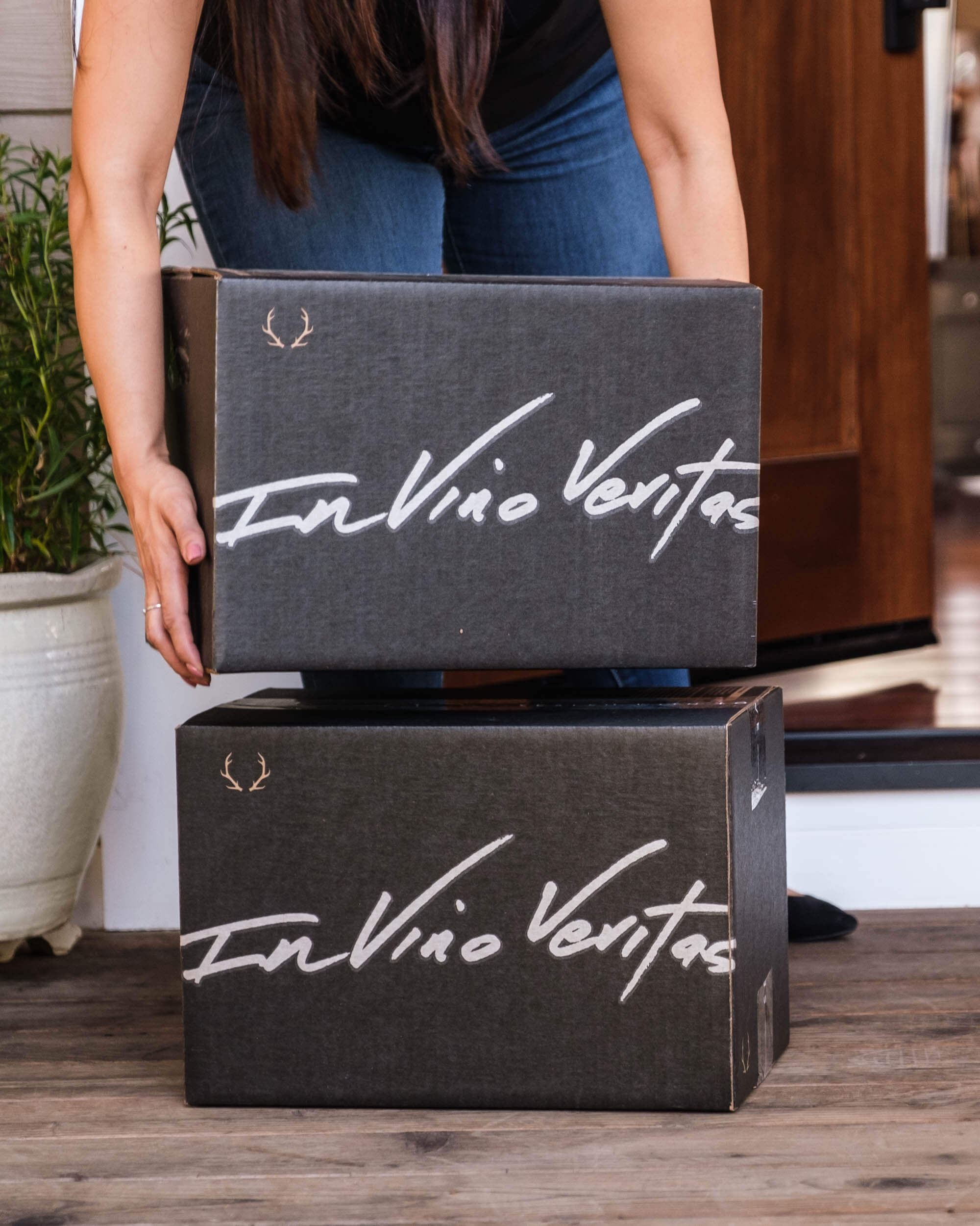 A woman picks up a box from Dry Farm Wines off a porch. The boxes read "In Vino Veritas" 