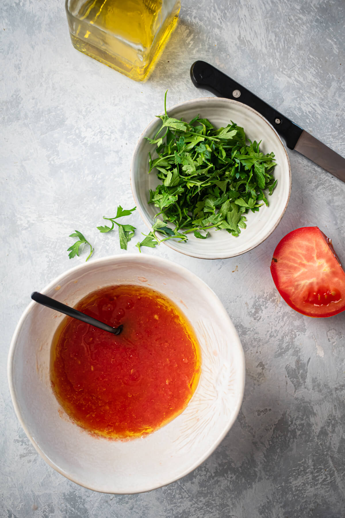 A bowl of tomato pulp with olive oil sits on a grey countertop next to another bowl of parsley. 