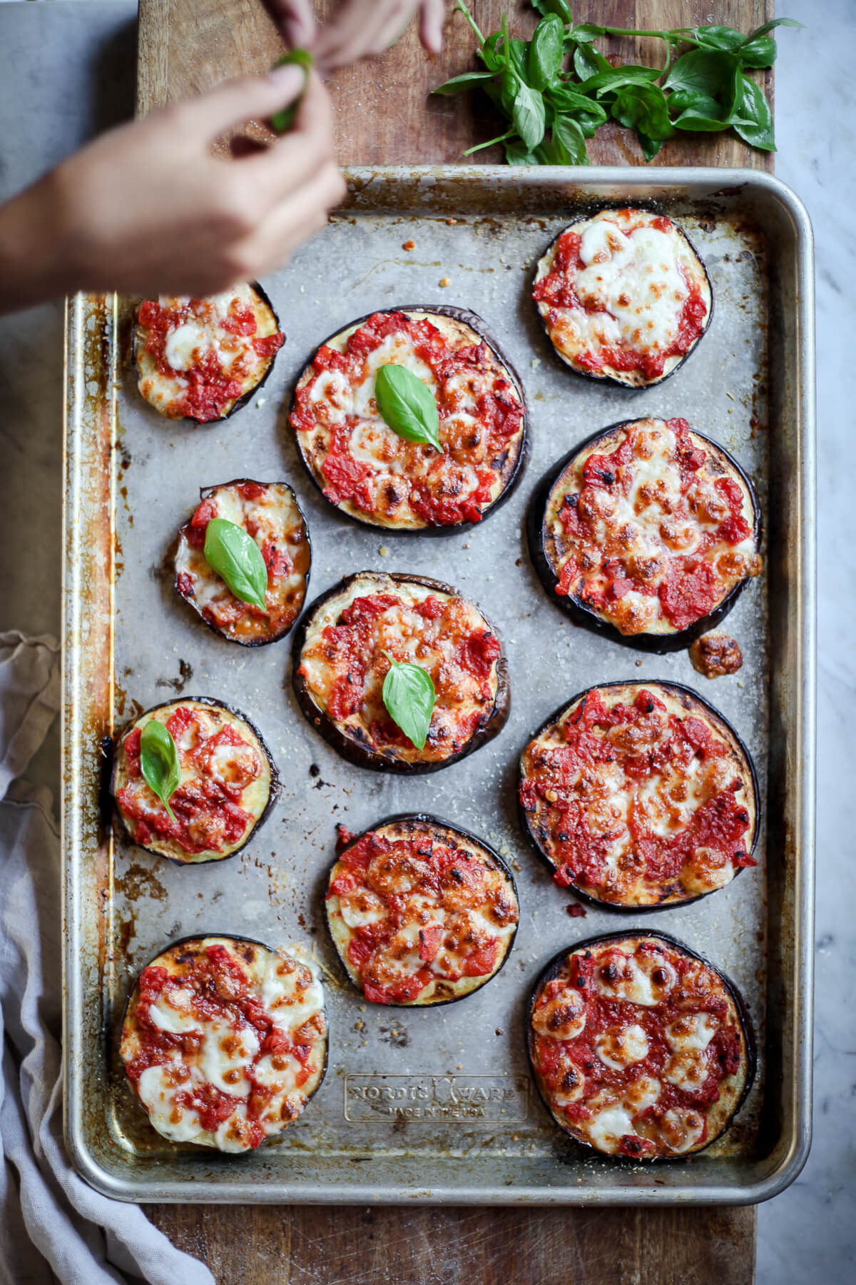 An overhead photo of Julia Child's eggplant pizza bites on a baking sheet while hands top them with fresh basil. 
