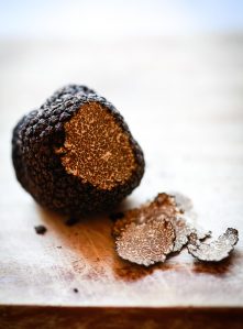 A fresh black truffle sits on a wooden cutting board. A few thin pieces have been shaved off to reveal a marbled interior.