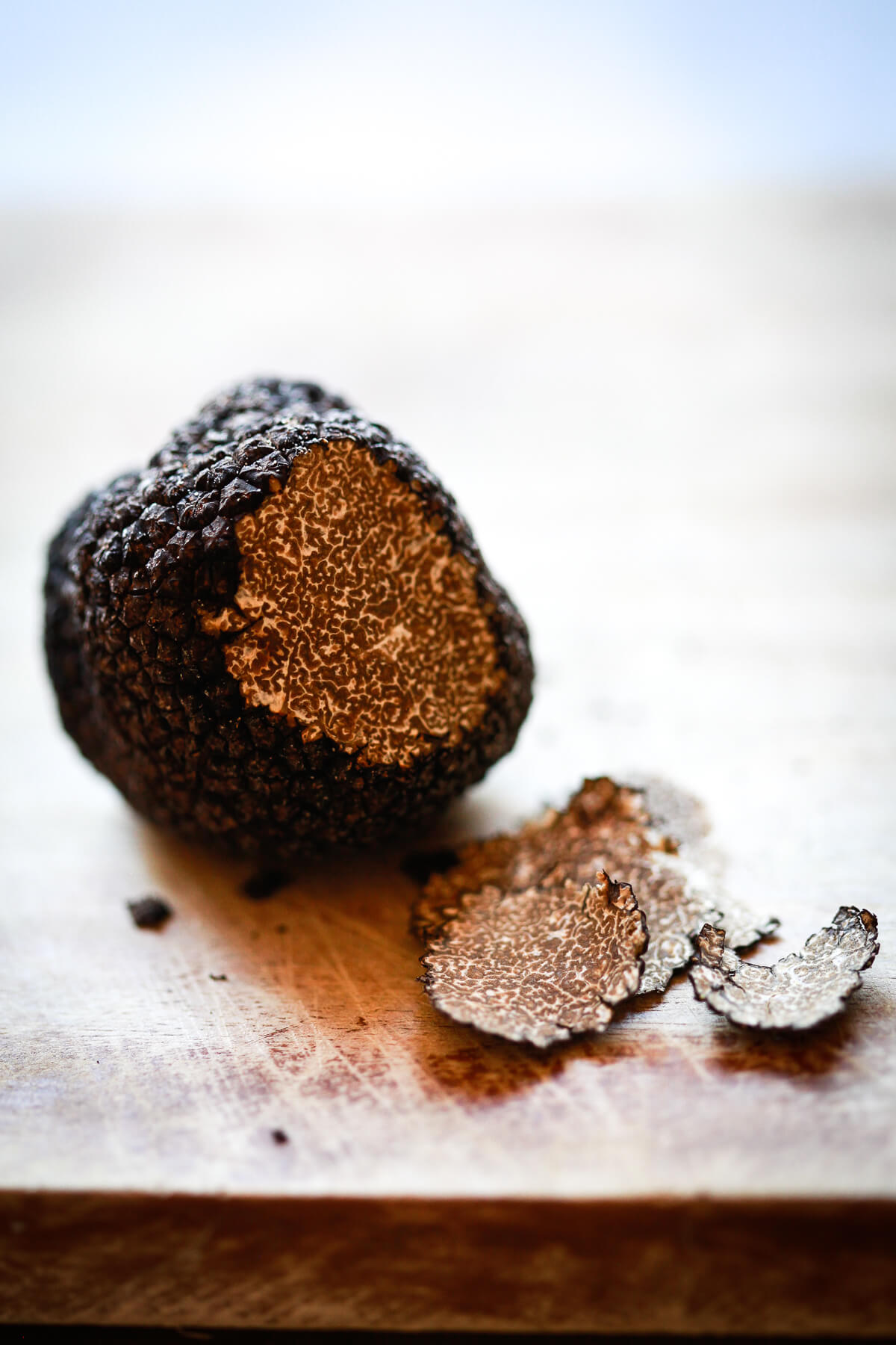 A fresh black truffle sits on a wooden cutting board. A few thin pieces have been shaved off to reveal a marbled interior. 