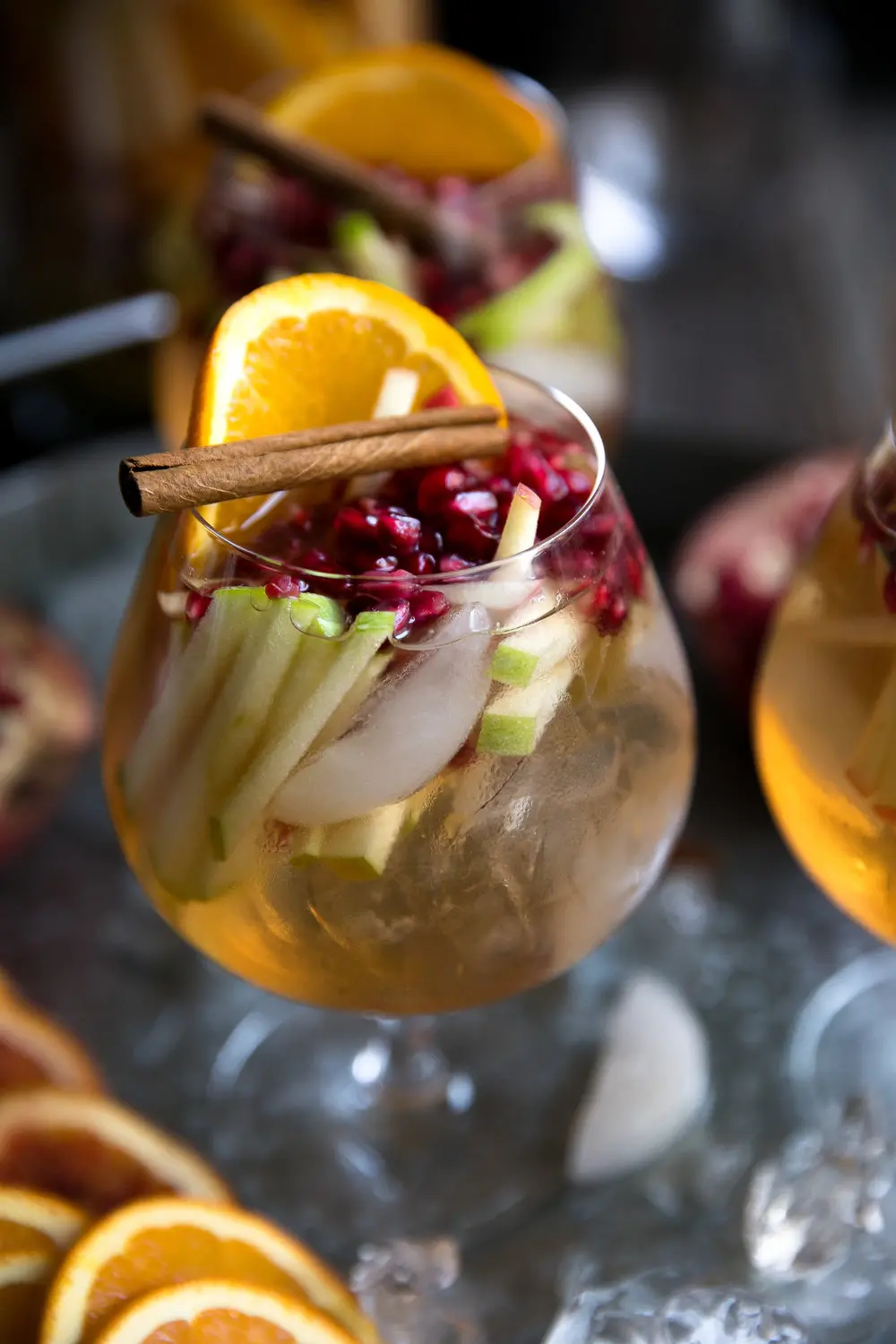 A clear wine glass filled with apple cider sangria garnished with pomegranate arils, sliced apples, orange, and a cinnamon stick. 