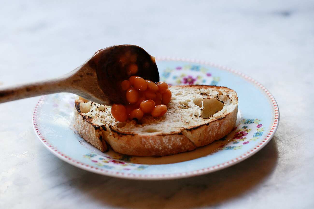 Heinz beans in tomato sauce is spooned onto a slice of toast with a wooden spoon.