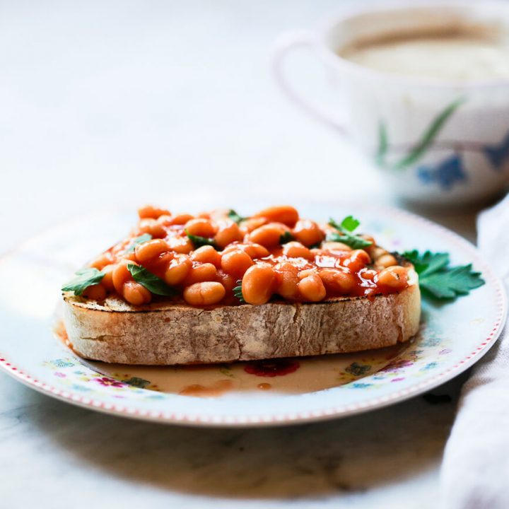 A thick slice of rustic toast topped with baked beans sits on a small plate in a white kitchen. This is classic British beans on toast recipe.