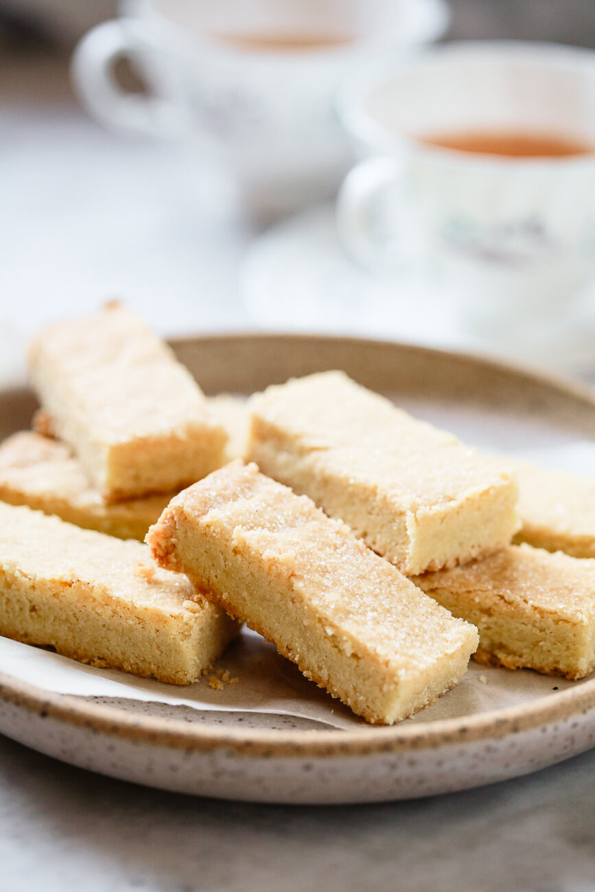 A beautiful photo of traditional Scottish shortbread (like Walker's shortbread) cookies on a ceramic plate with tea cups in the background. 