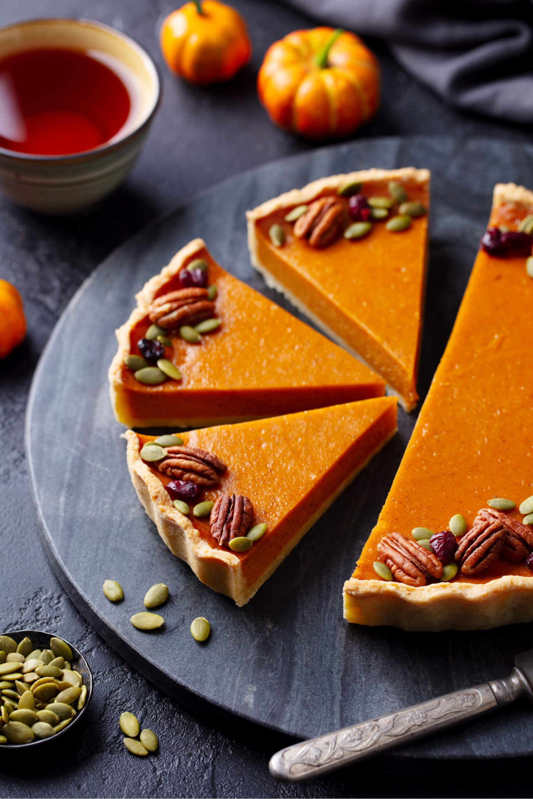 A pumpkin pie is decorated with pecans and pumpkin seeds. The pie is cut into slices and sits on a slate tray. 