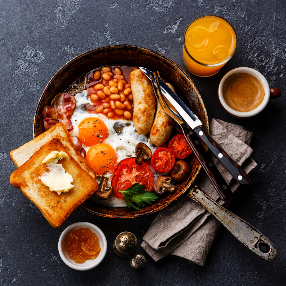An overhead photo of a full English breakfast including beans, toast, eggs, tomatoes, and sausage served in a skillet. 