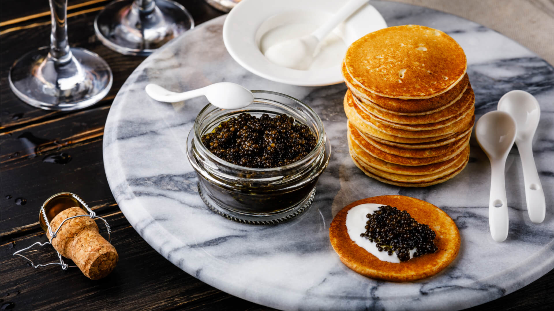 A glass bowl filled with black caviar and a white spoon sits on a marble platter next to blini Russian pancakes. 