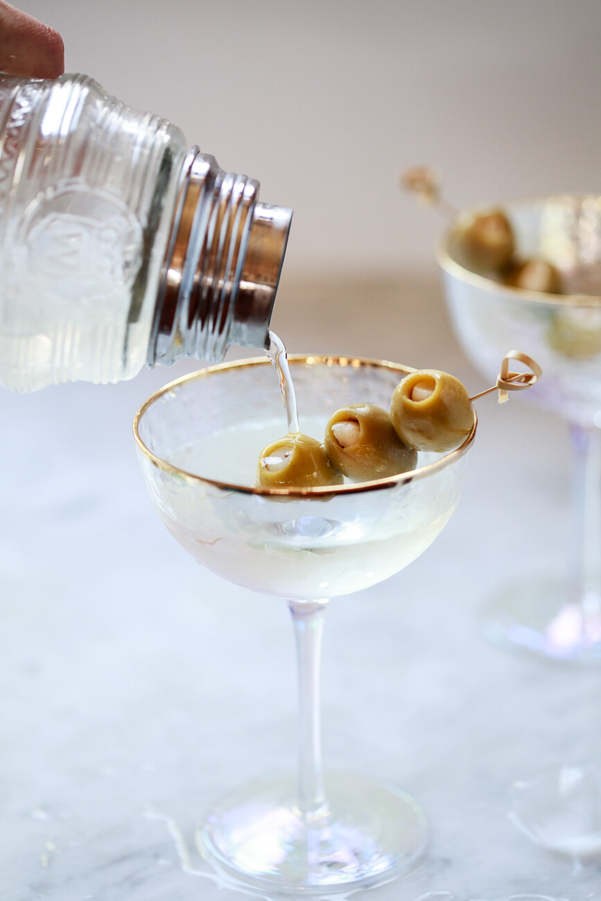 A homemade martini is poured from a cocktail shaker into a beautiful coupe glass filled with skewered olives.