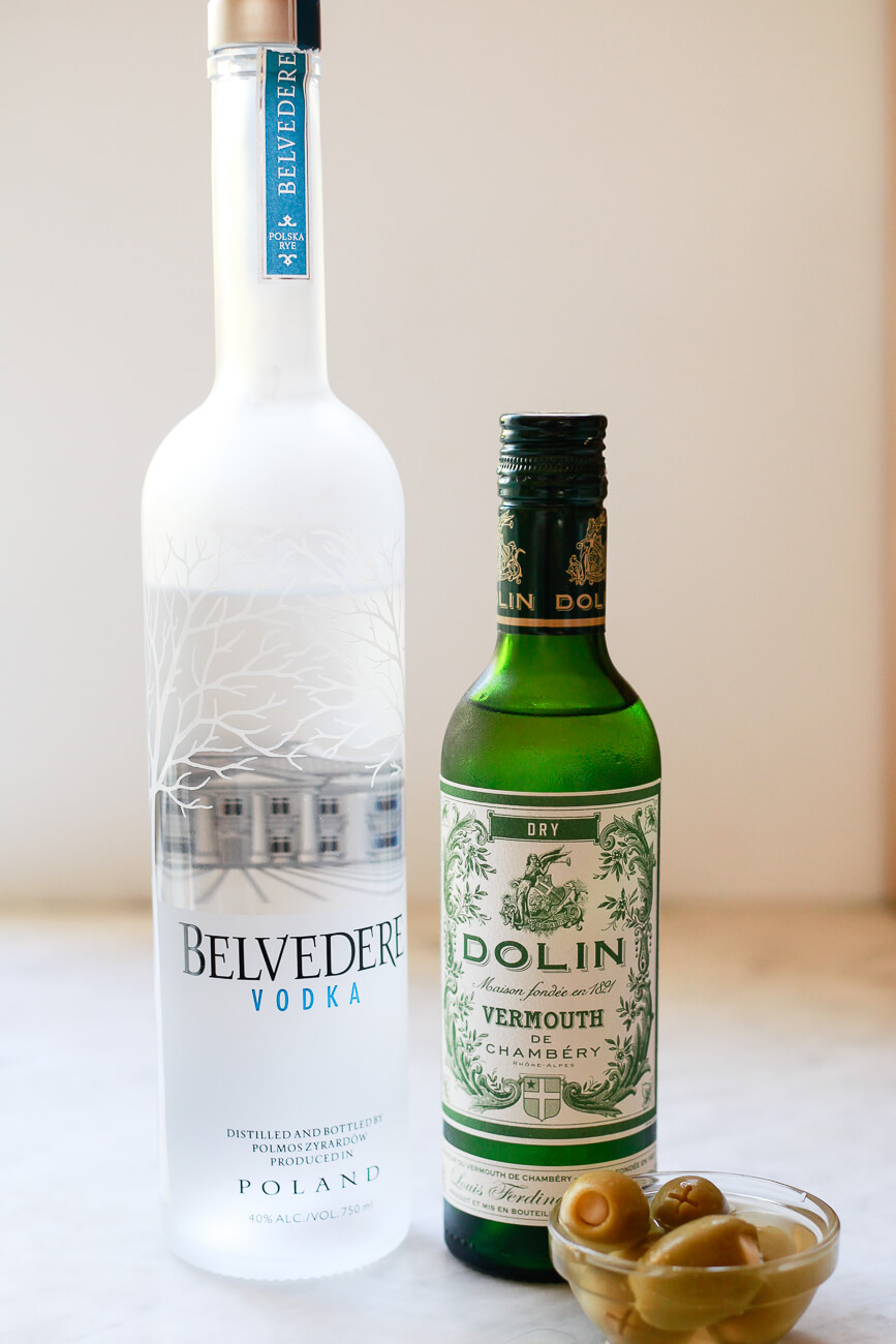 Bottles of vodka and dry white vermouth sit on a marble countertop next to green olives. 