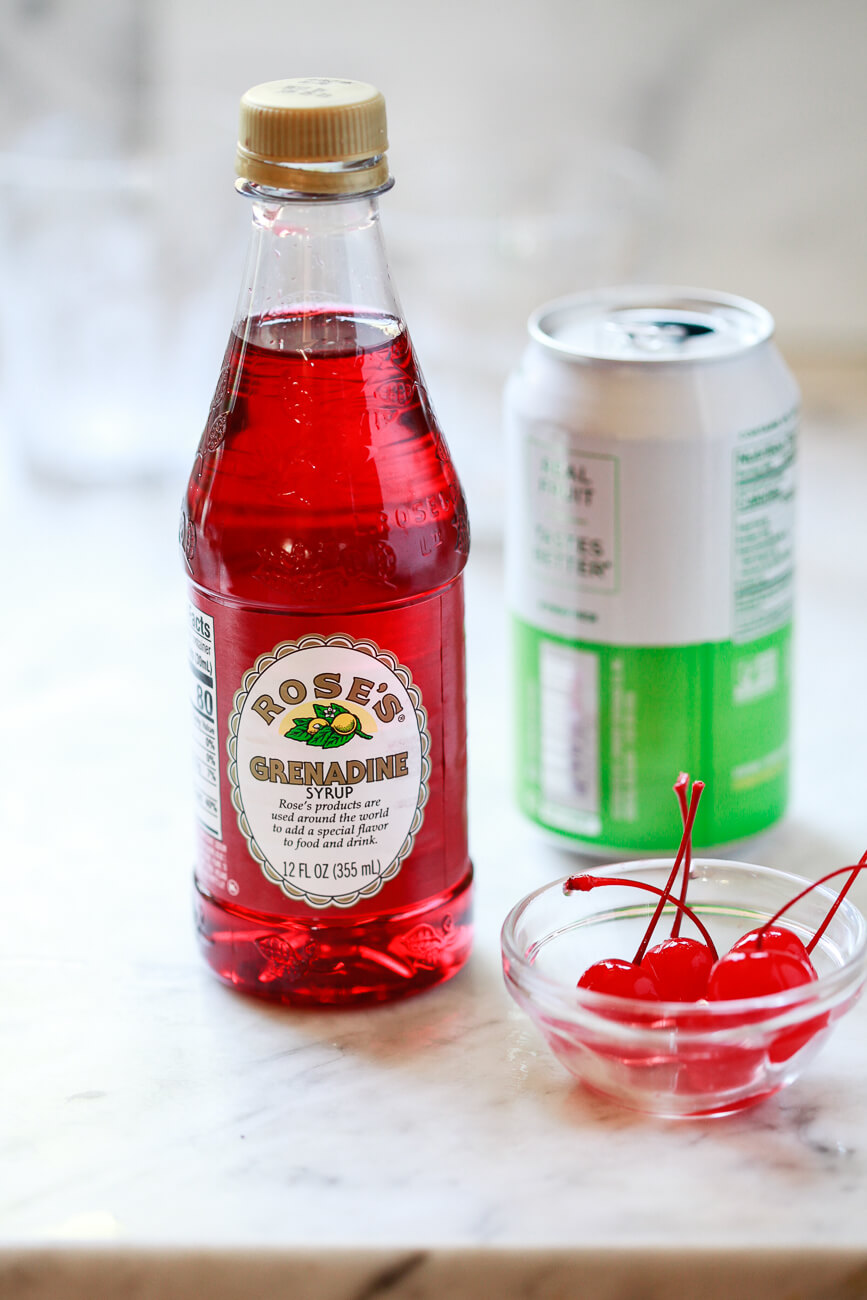 The ingredients for a Shirley Temple recipe sit on a marble countertop: Rose's grenadine syrup, a can of lemon lime soda, and maraschino cherries. 