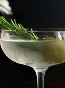 A close-up photo of a cold dirty martini in a coupe glass with a sprig of rosemary and a green olive.