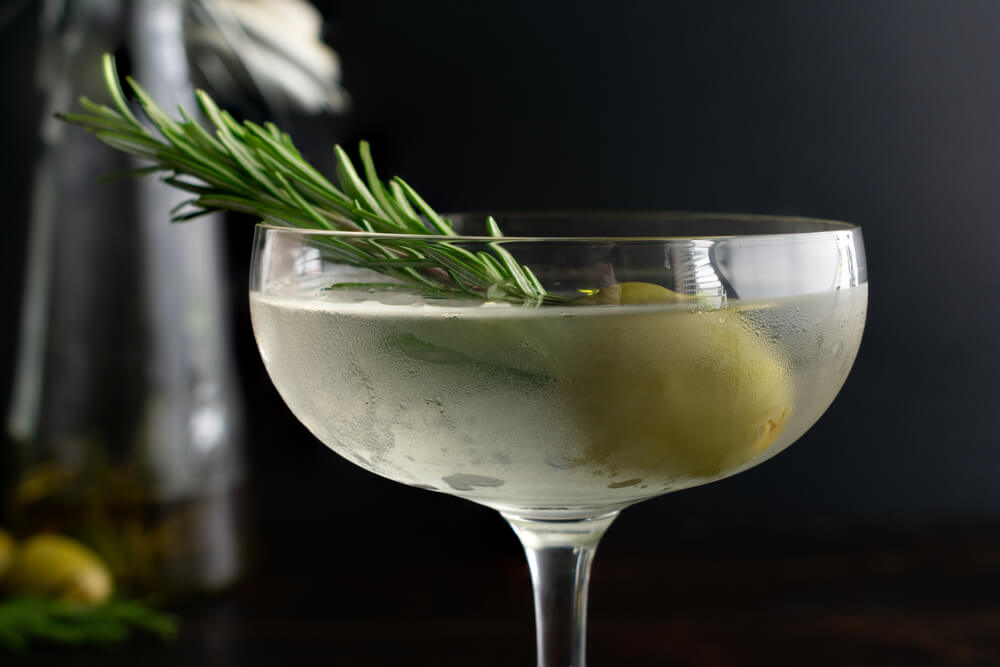 A close-up photo of a cold dirty martini in a coupe glass with a sprig of rosemary and a green olive. 