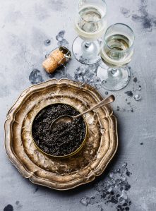 A beautiful overhead photo of caviar on a bowl of crushed ice on a silver platter. Two glasses of Champagne sit on the side.