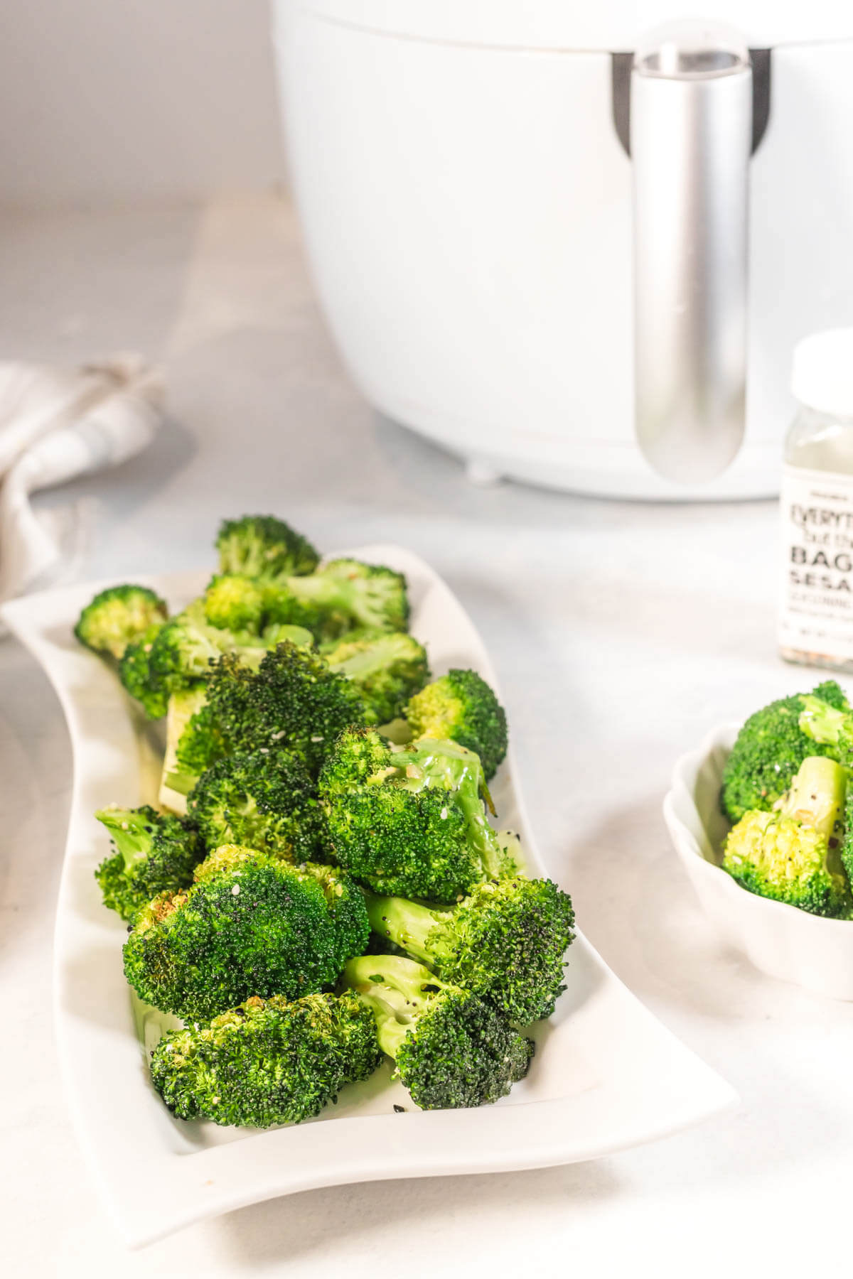 A white dish of air fryer broccoli florets on a white counter with a white air fryer in the background. 