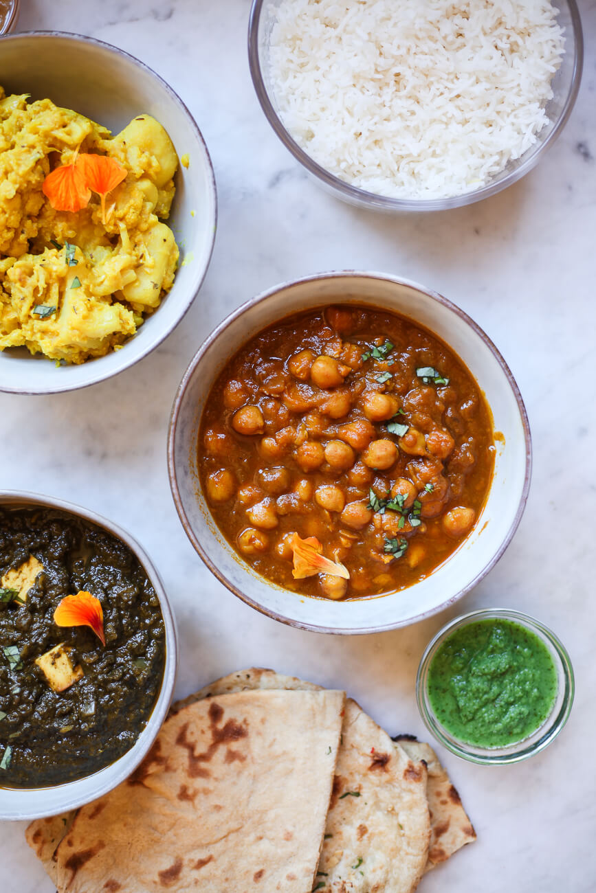 Ceramic bowls filled with vegetarian Indian dishes. Clockwise, aloo gobi, basmati rice, chana masala, mint chutney, chapati, and saag. 