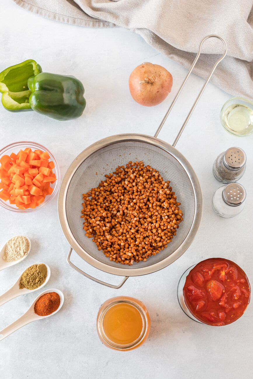 All the ingredients for lentil chili sit on a white countertop: green bell pepper, onion, salt and pepper, diced carrot, spices, vegetable broth, lentils, and tomatoes. 