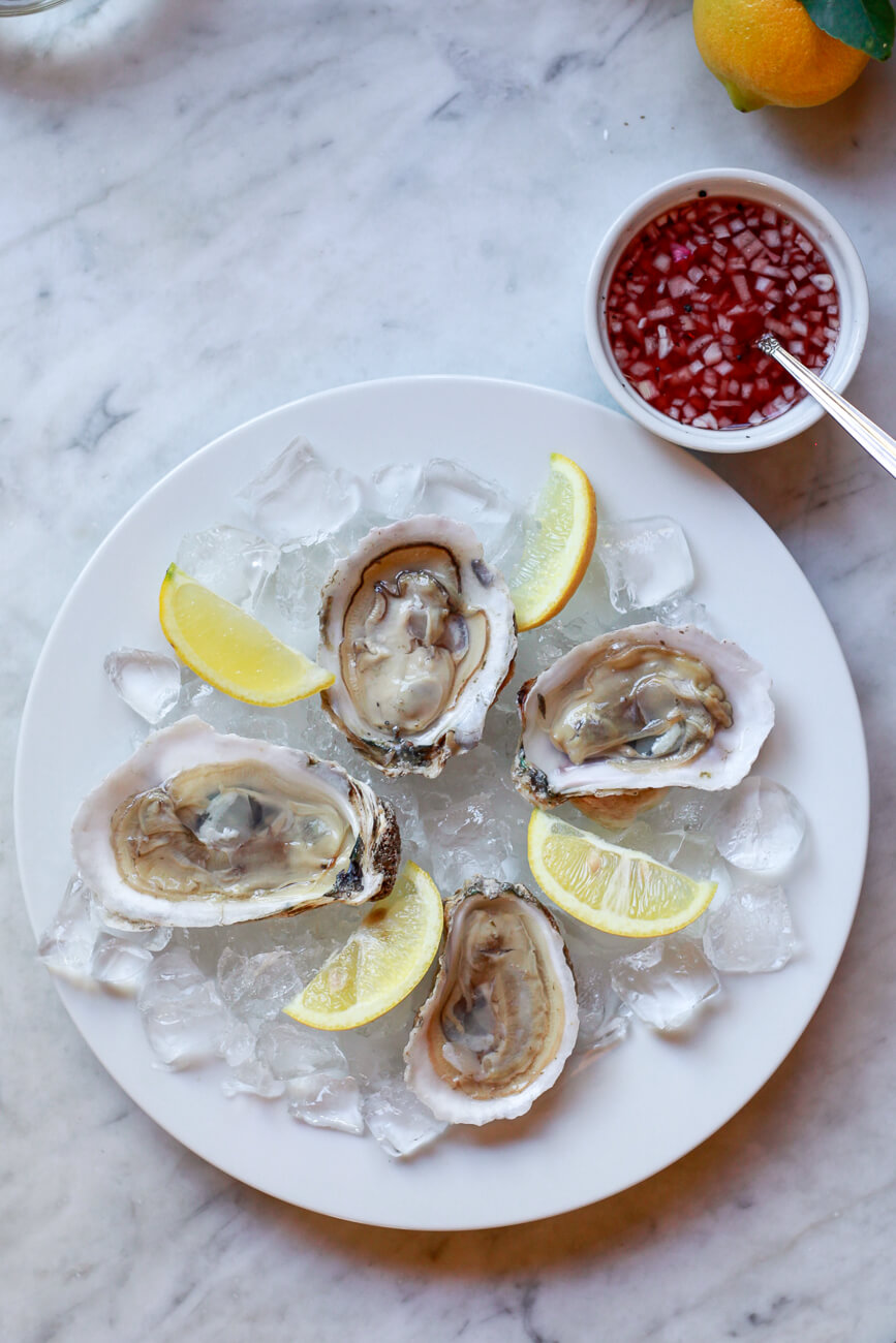 4 raw oysters on the half shell on a plate of ice with lemon wedges and a bowl of mignonette sauce on the side. 