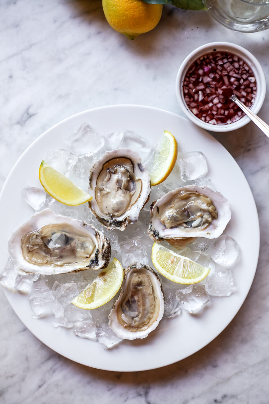 Raw oysters on the half shell on ice with lemon slices and a side of mignonette sauce on a marble countertop as an expensive of the most expensive foods in the world. 