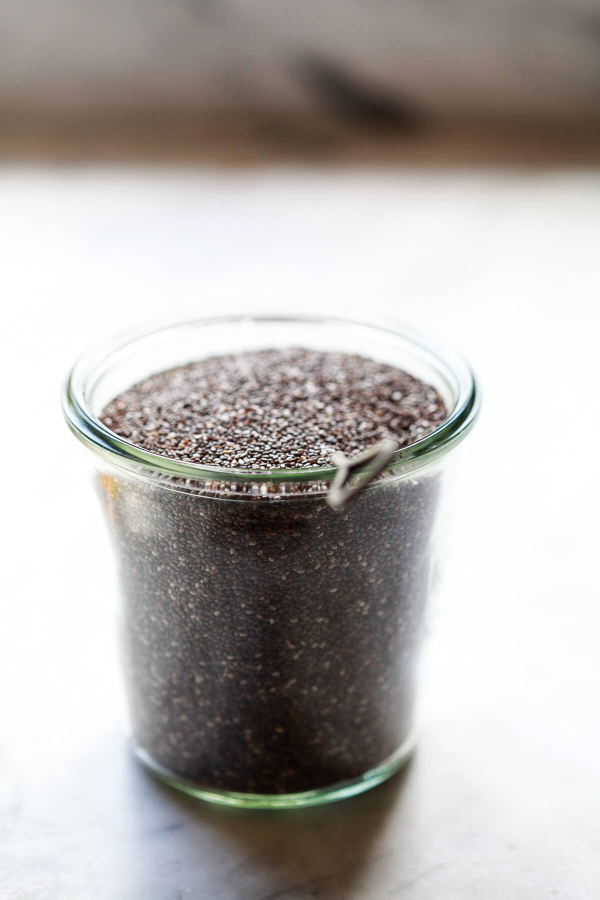A Weck jar filled with chia seeds sits on a marble countertop with a metal spoon. 